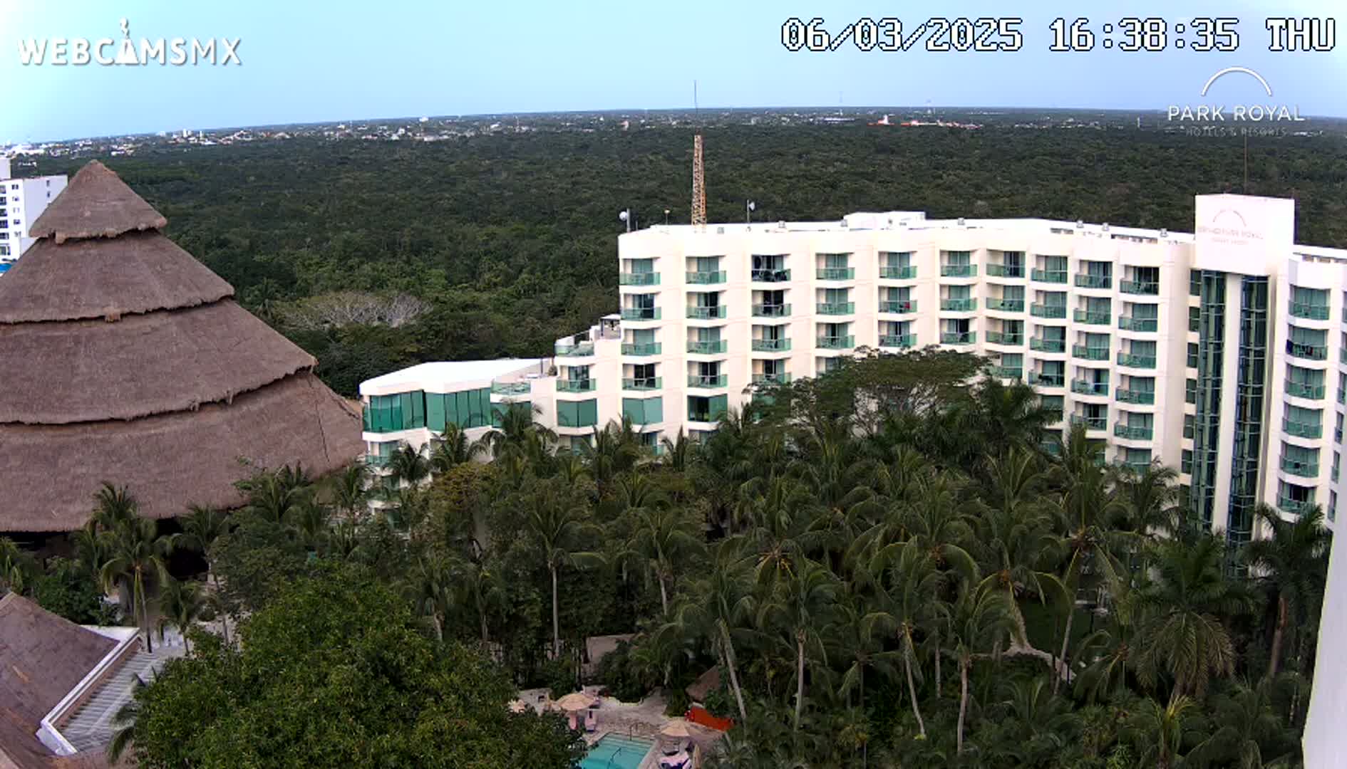 An aerial view shows a large, multi-story hotel nestled among lush palm trees and tropical vegetation under a clear sky.
