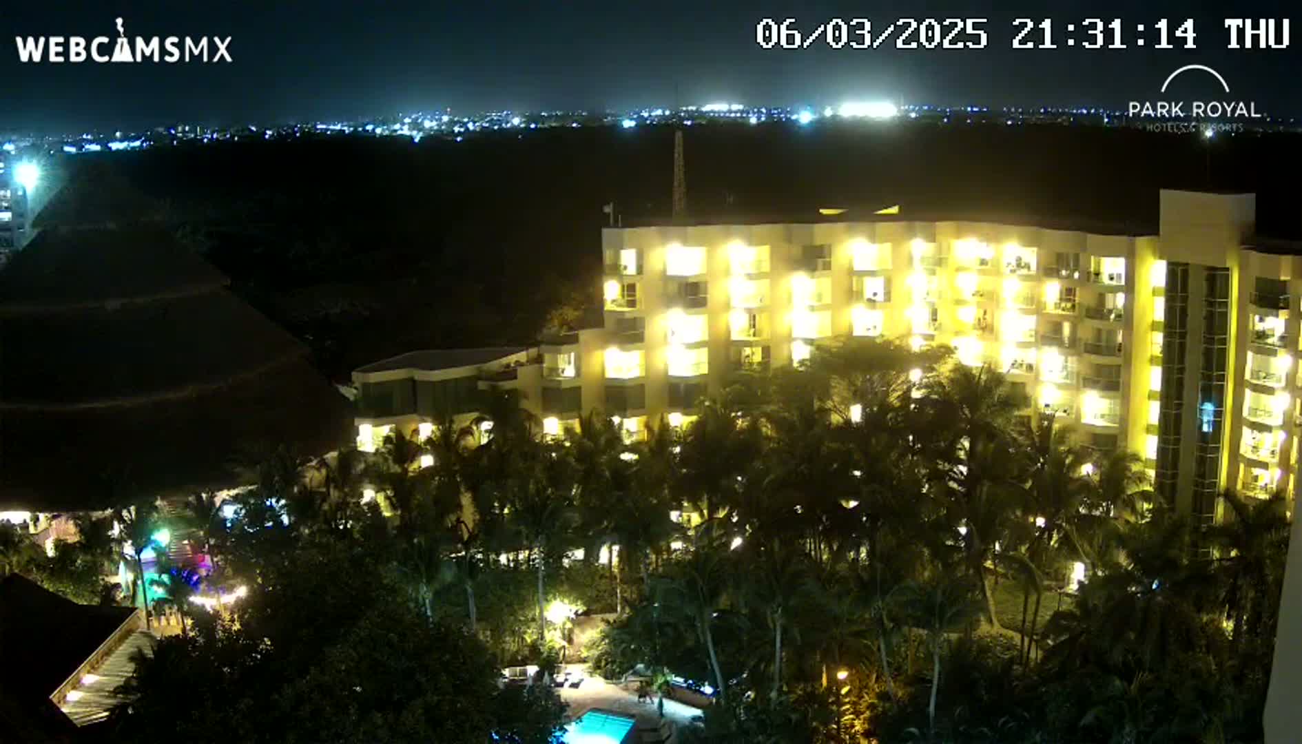 A brightly lit, multi-story hotel at night is surrounded by palm trees and a swimming pool, under a clear, dark sky with distant city lights visible in the background.