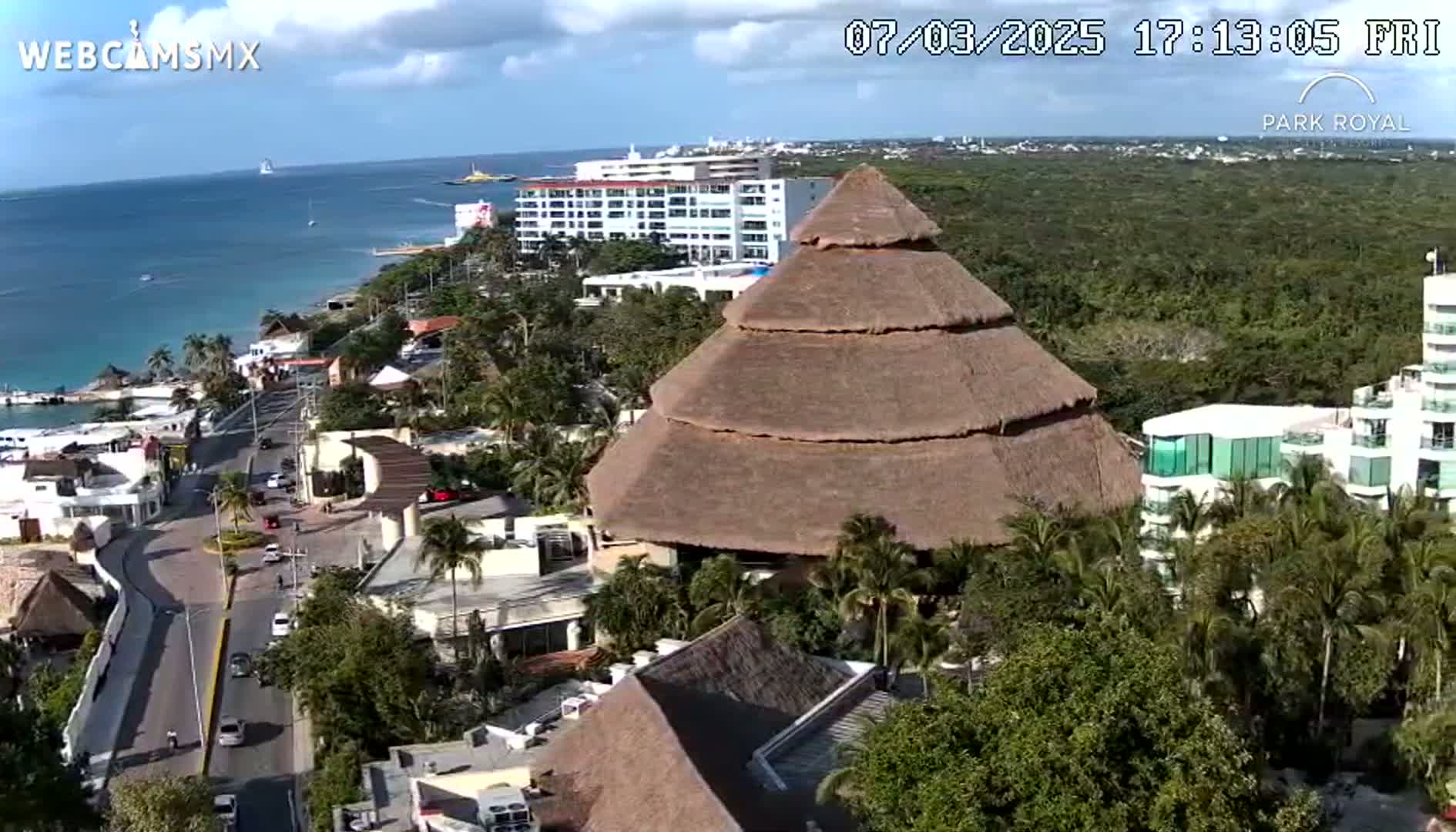 A coastal scene on a partly cloudy day shows a large thatched-roof building near a road and hotels, with a body of water and distant coastline visible in the background.