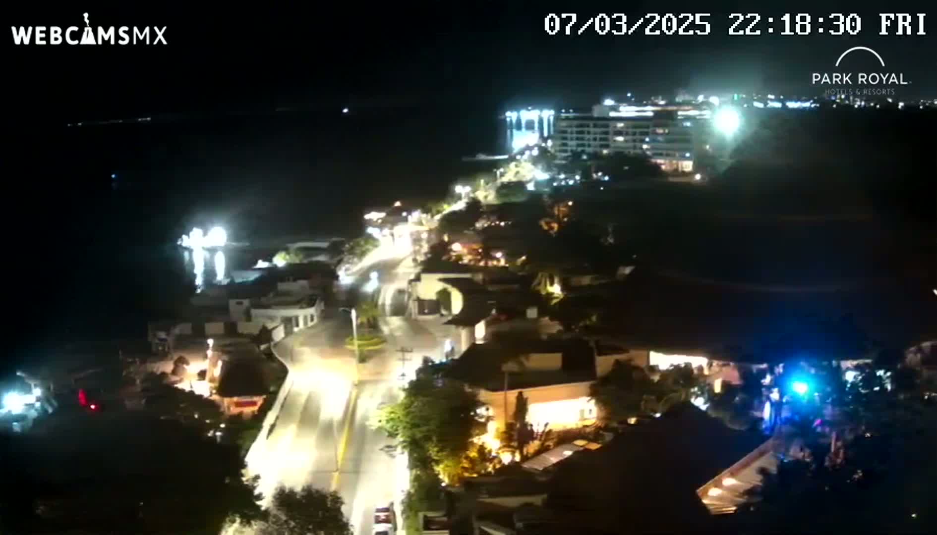 A nighttime aerial view of a coastal town shows a road winding through buildings near a body of water, with a large hotel illuminated in the background under a clear, dark sky.