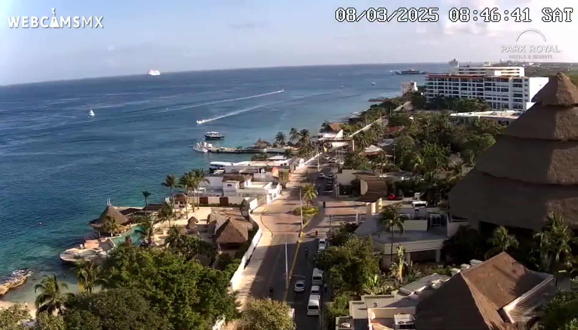 An aerial view shows a coastal town on a sunny day, with a clear blue ocean featuring boats and a cruise ship in the distance, and a road leading to a large resort.