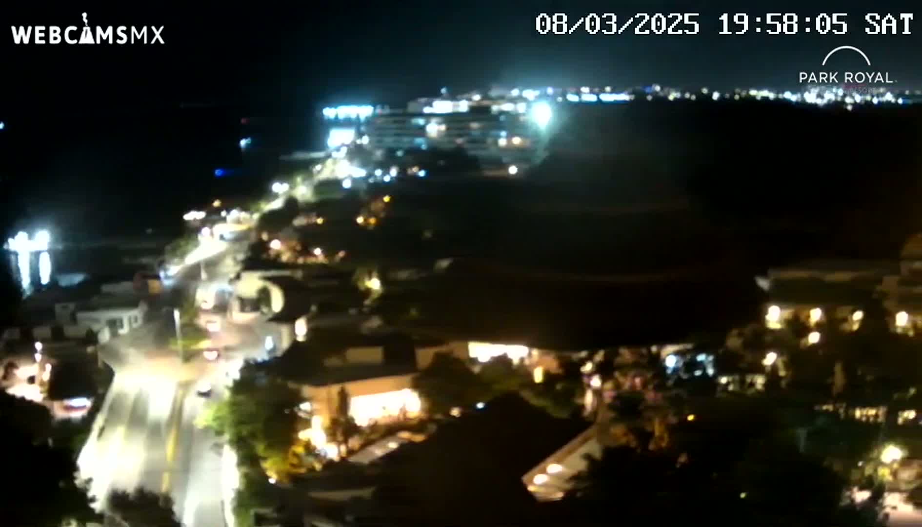 A nighttime, high-angle view shows a coastal town with illuminated buildings and roads, and a body of water in the background.