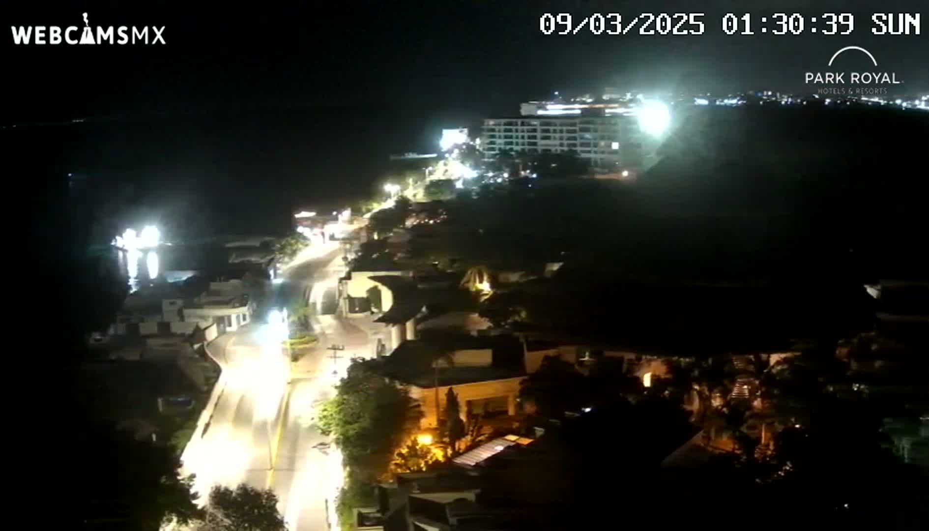 A nighttime aerial view shows a coastal town with a roadway illuminated by streetlights, leading to a large hotel overlooking the dark ocean under a clear sky.