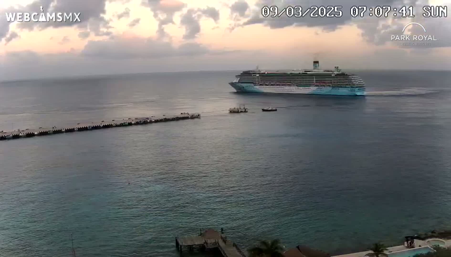 A large cruise ship is docked near a long pier in calm waters under a partly cloudy sunrise sky.