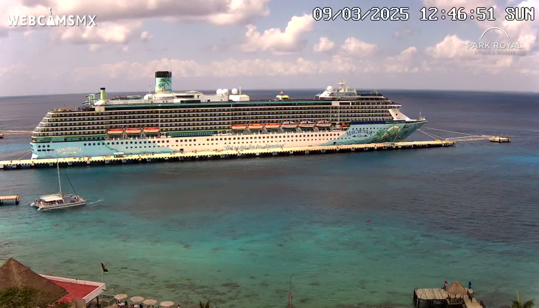 A large cruise ship is docked at a pier on a partly cloudy day with calm, turquoise water.