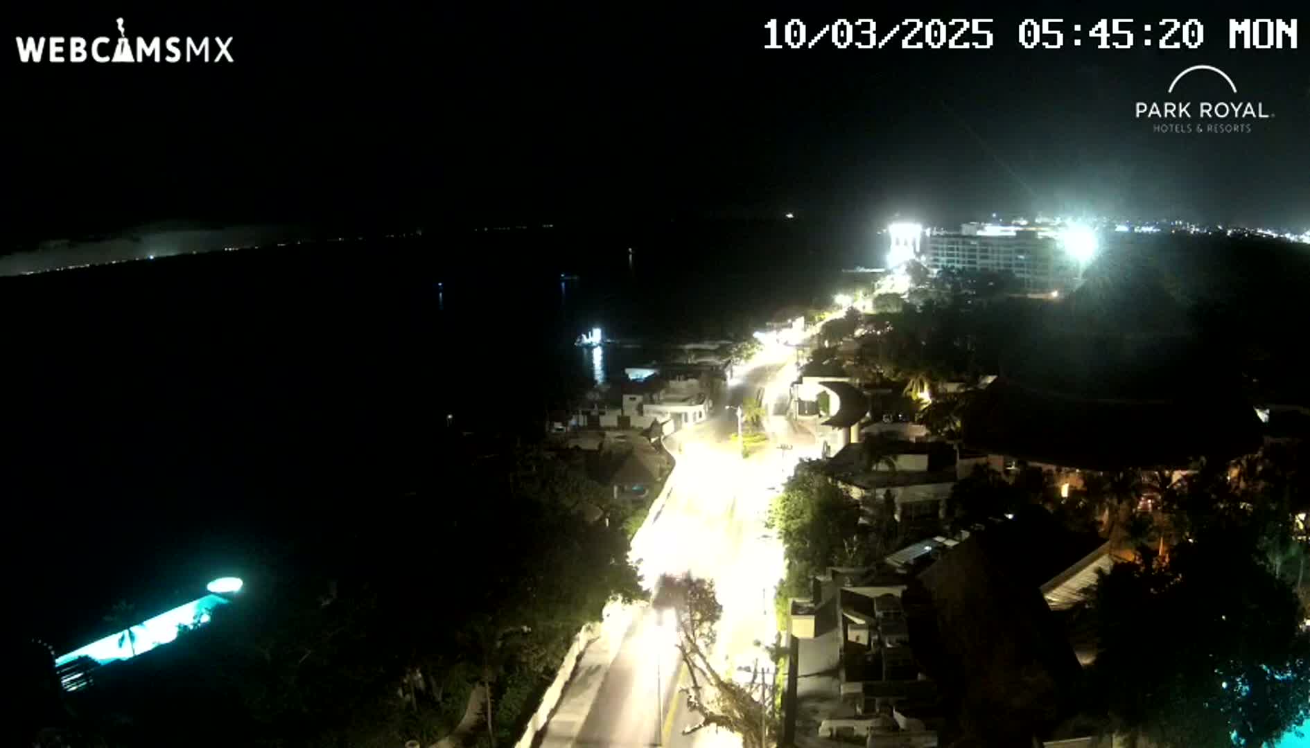 A nighttime, aerial view of a coastal road illuminated by streetlights, leading to a brightly lit resort complex near a dark body of water.