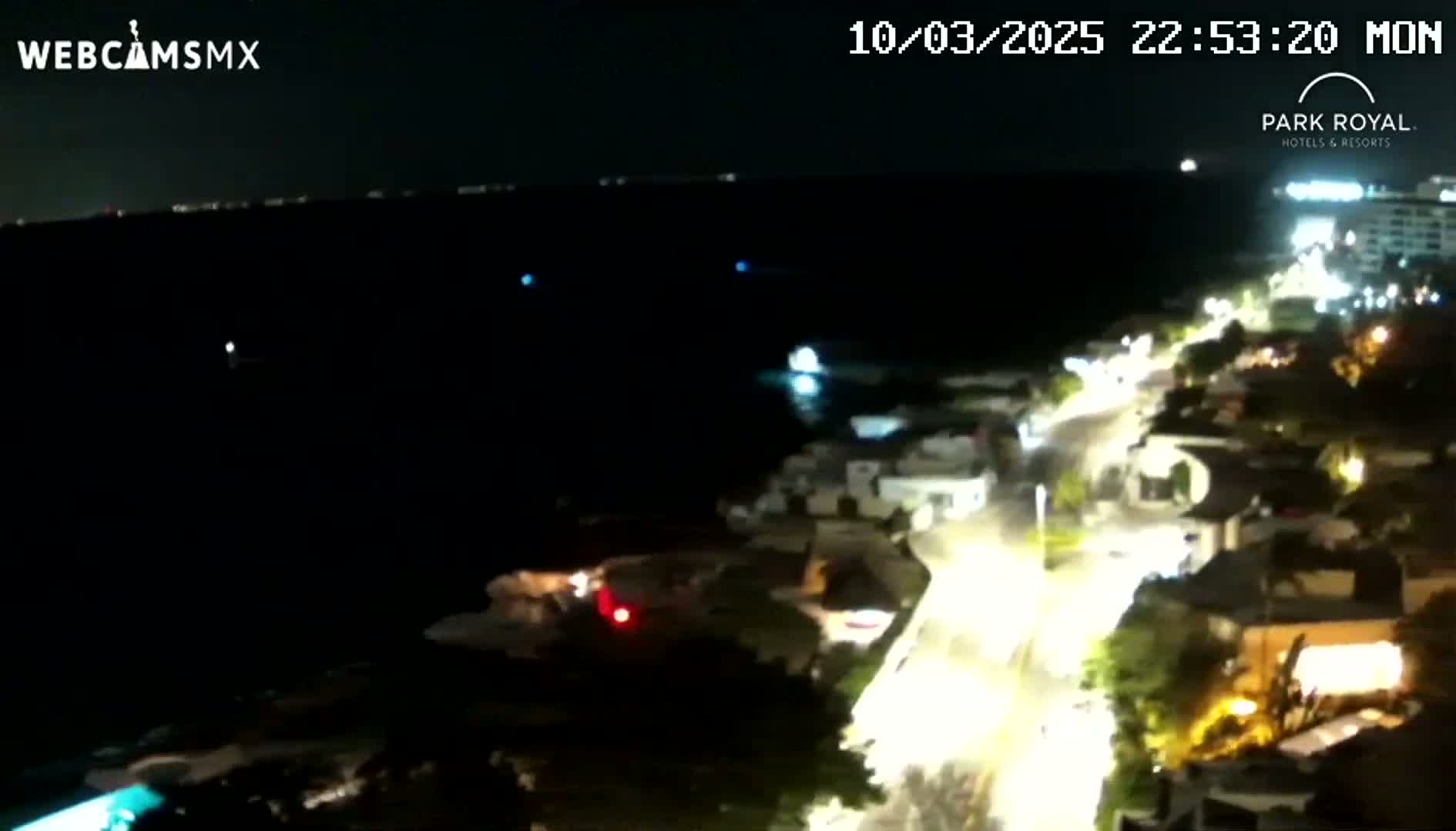 A nighttime coastal scene shows a dark body of water, a brightly lit road curving along a shoreline lined with buildings.