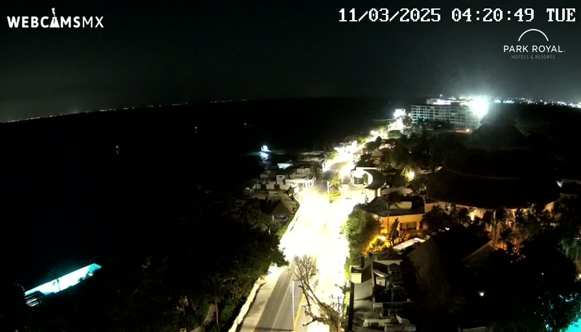 A nighttime aerial view shows a brightly lit road winding through a coastal town next to a dark ocean under a clear sky.