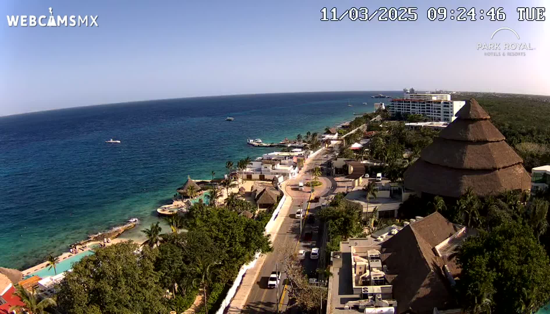 An aerial view of a coastal resort town on a sunny day, showing a clear blue ocean, a road lined with buildings, and a large thatched-roof structure.