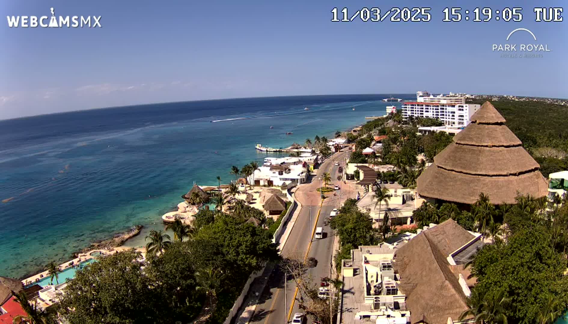 An aerial view shows a coastal resort town on a sunny day with clear blue water and a road winding through the buildings towards the ocean.