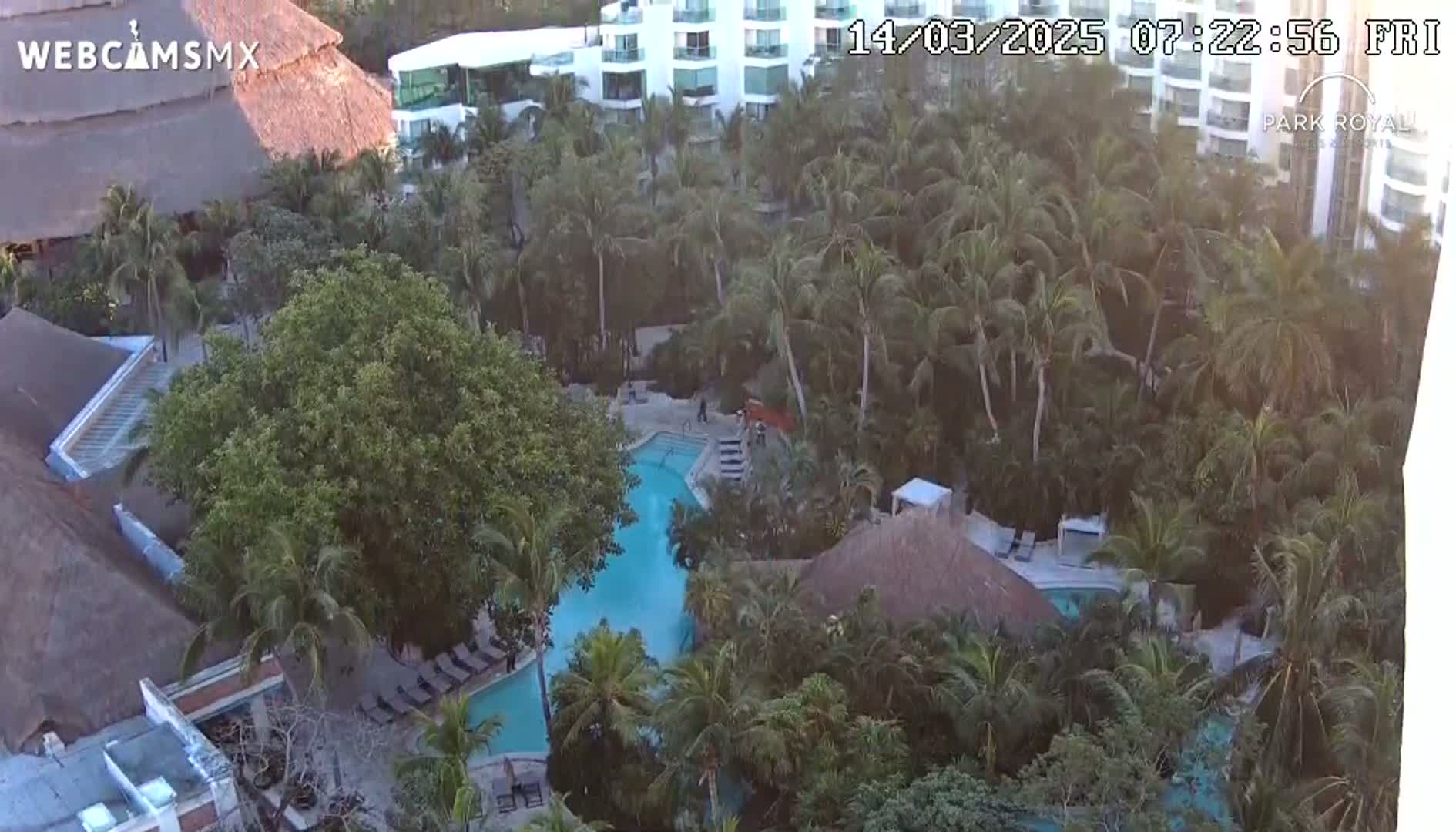 An aerial view shows a resort with multiple pools surrounded by lush tropical vegetation and buildings under a clear sky.