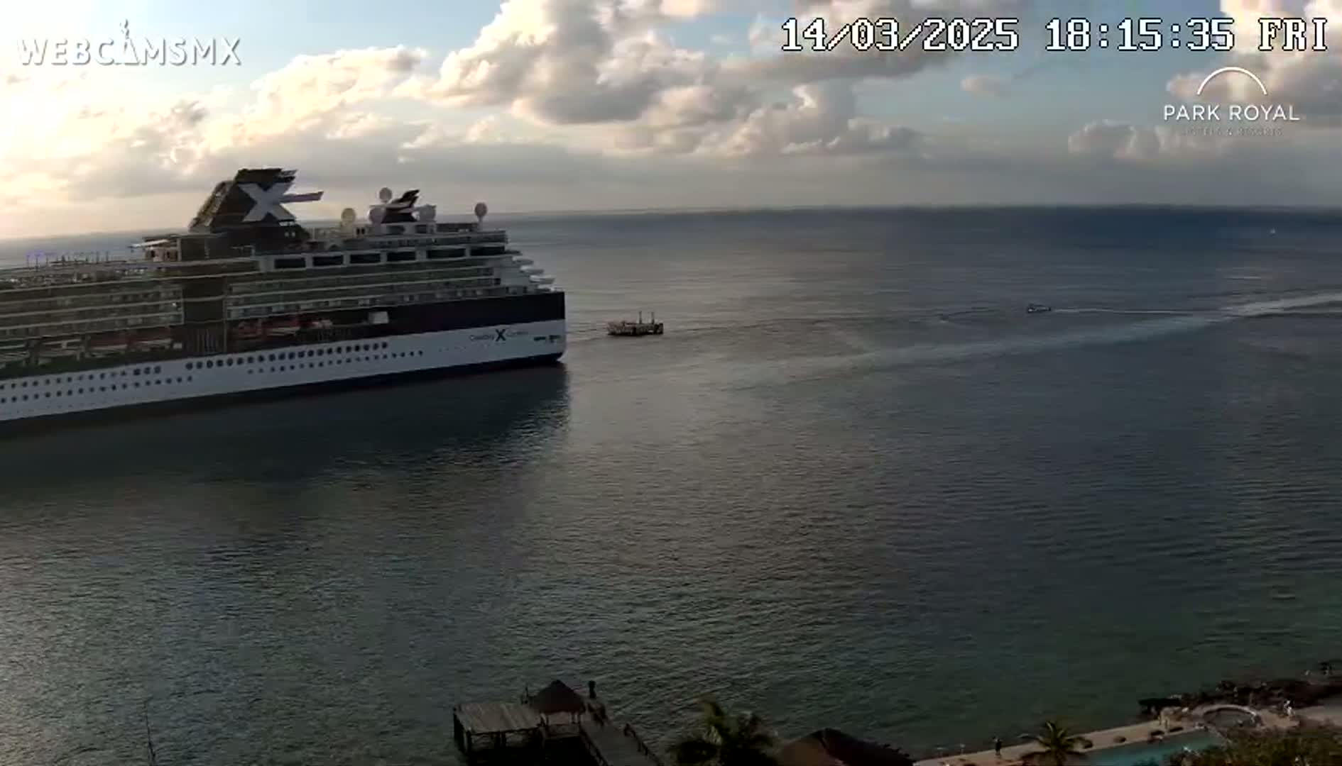 A large cruise ship is docked near a smaller boat in calm water under a partly cloudy sky.