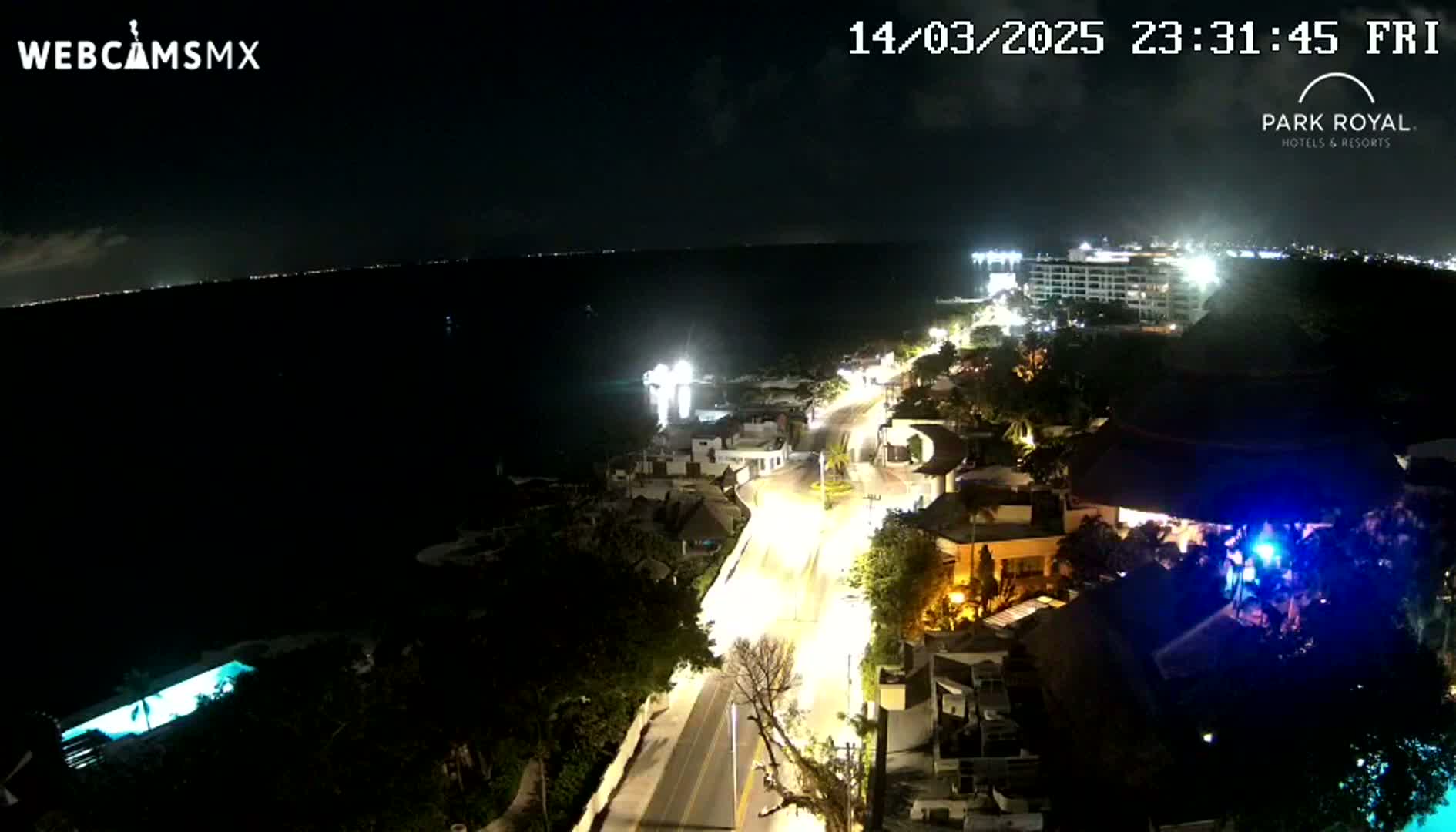 A nighttime aerial view shows a coastal town with illuminated buildings and streets near a dark body of water under a clear sky.