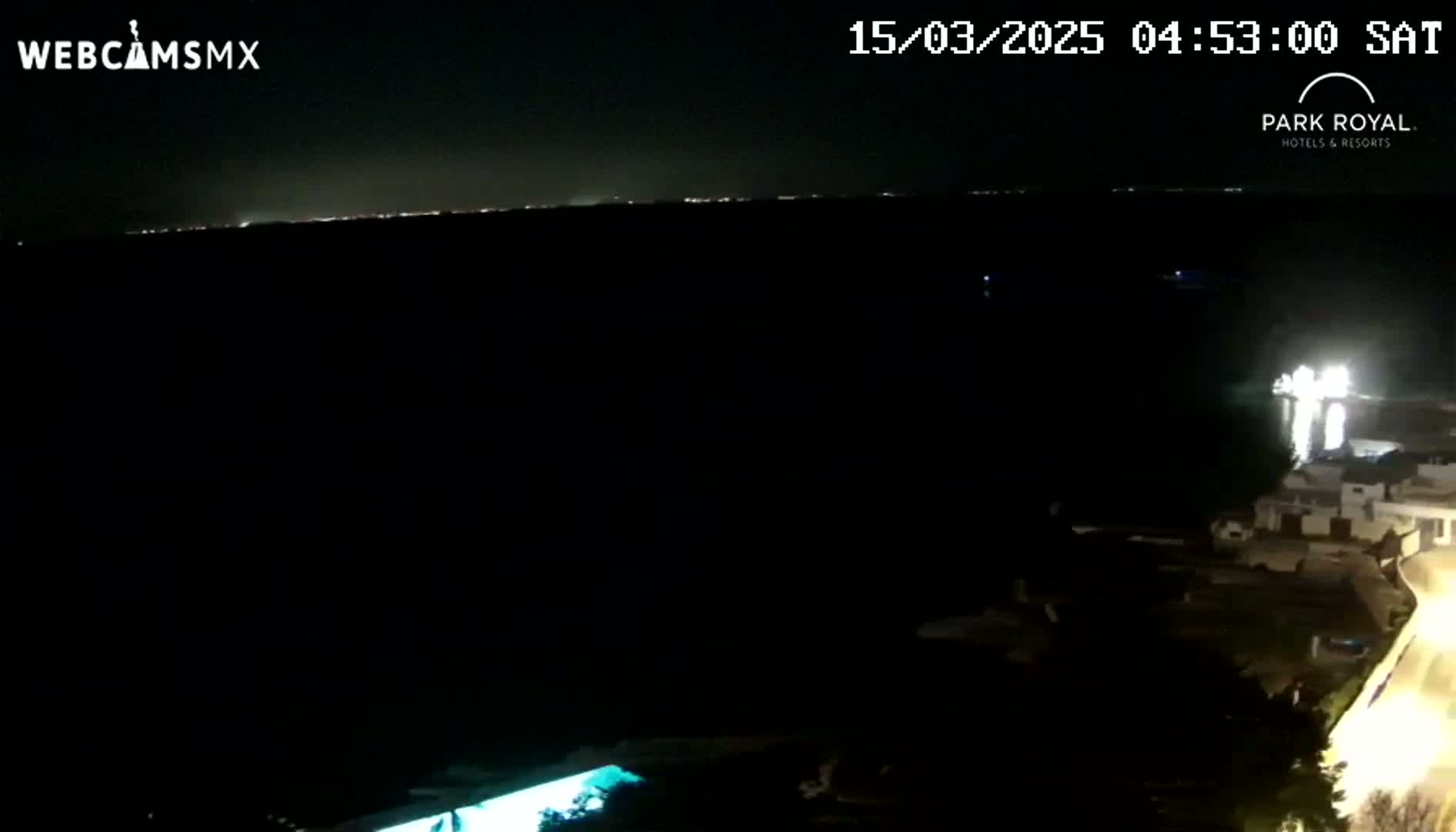 A nighttime, long-range view shows a dark body of water with a distant, faintly lit shoreline, and brightly lit buildings in the foreground to the right.
