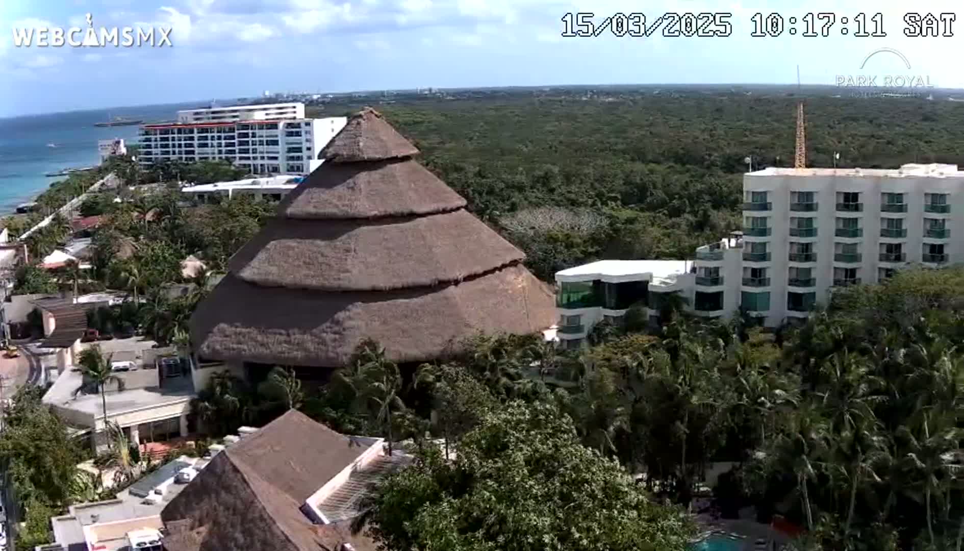 A large, tiered, thatched-roof structure is situated among numerous buildings and lush tropical vegetation near a coastline under partly cloudy skies.