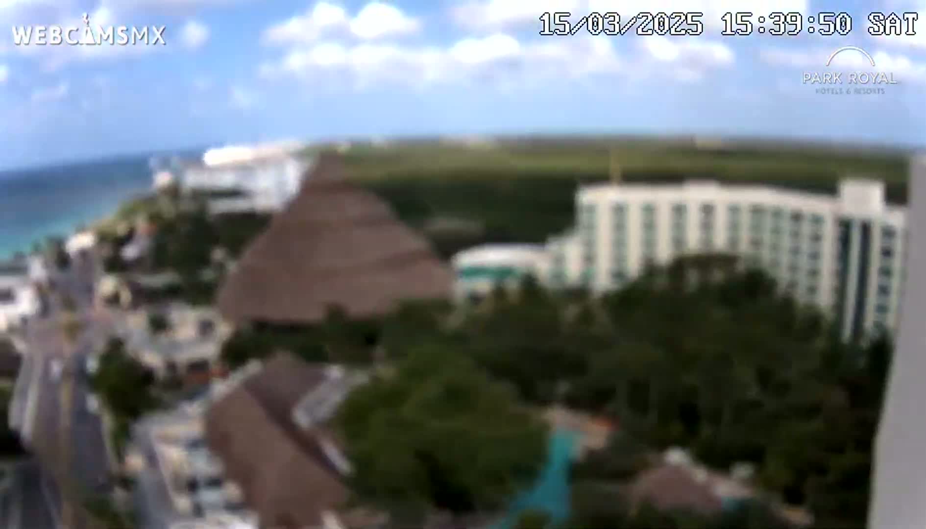 An aerial view shows a resort area with buildings, vegetation, and a beach with blue water under partly cloudy skies.
