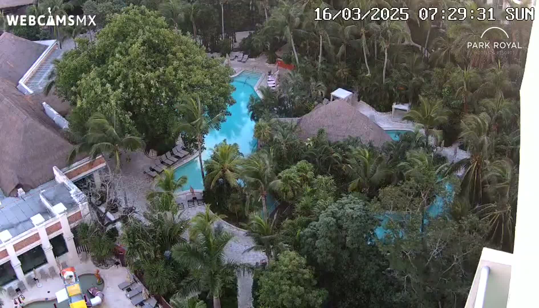 An aerial view shows a resort pool area with multiple pools surrounded by lush tropical vegetation under a sunny sky.