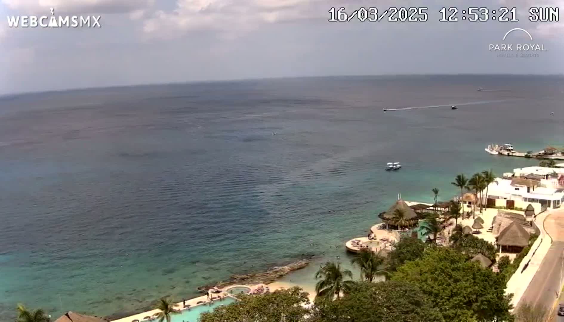 An aerial view shows a tropical resort on a coast with calm, clear blue water under a partly cloudy sky.