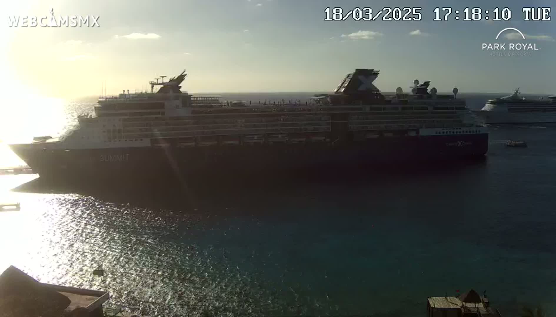 Two large cruise ships are docked in calm water under a mostly clear, sunny sky.