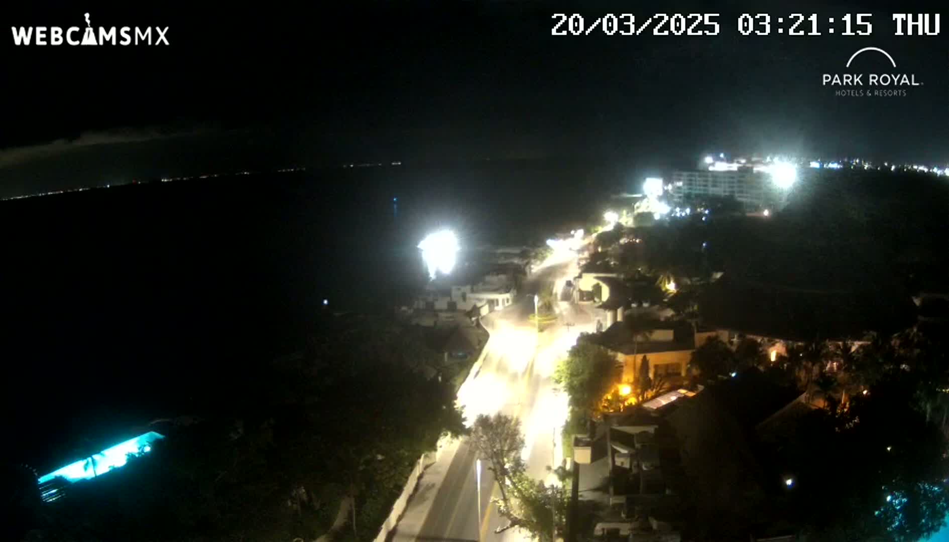 A nighttime, high-angle view of a coastal road with buildings and lights, overlooking a dark body of water under a clear sky.