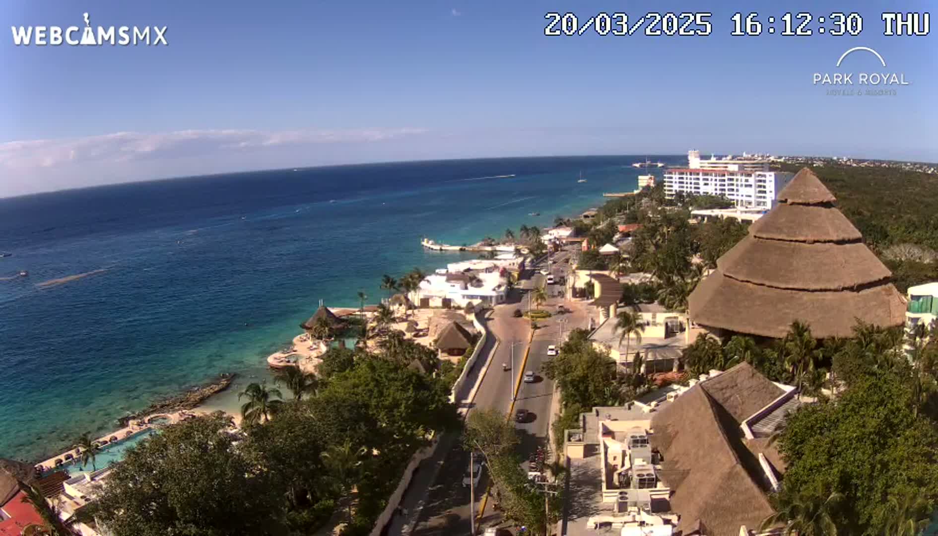 An aerial view shows a coastal resort town on a sunny day, with a turquoise ocean in the background and a large thatched-roof structure in the foreground.