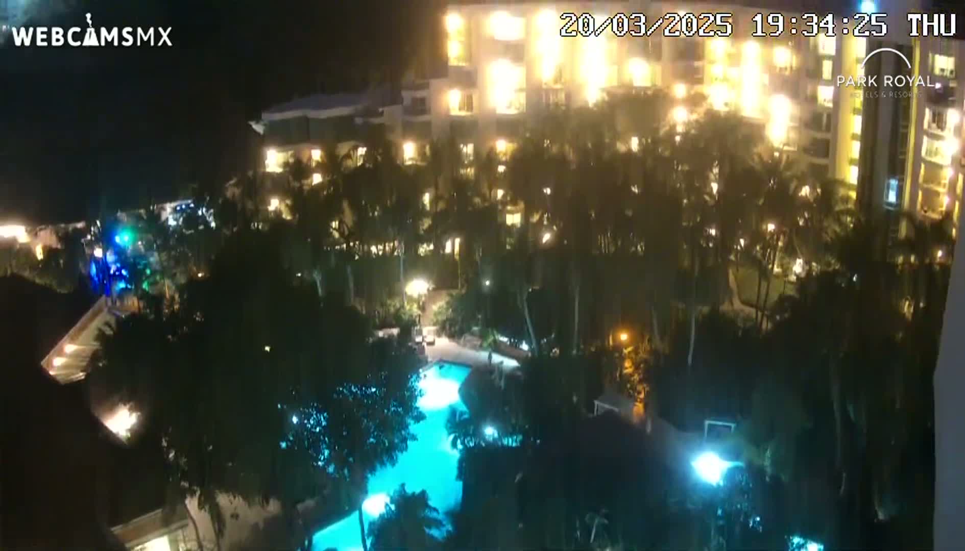 A nighttime aerial view of a resort with a lit swimming pool surrounded by palm trees and brightly lit buildings.
