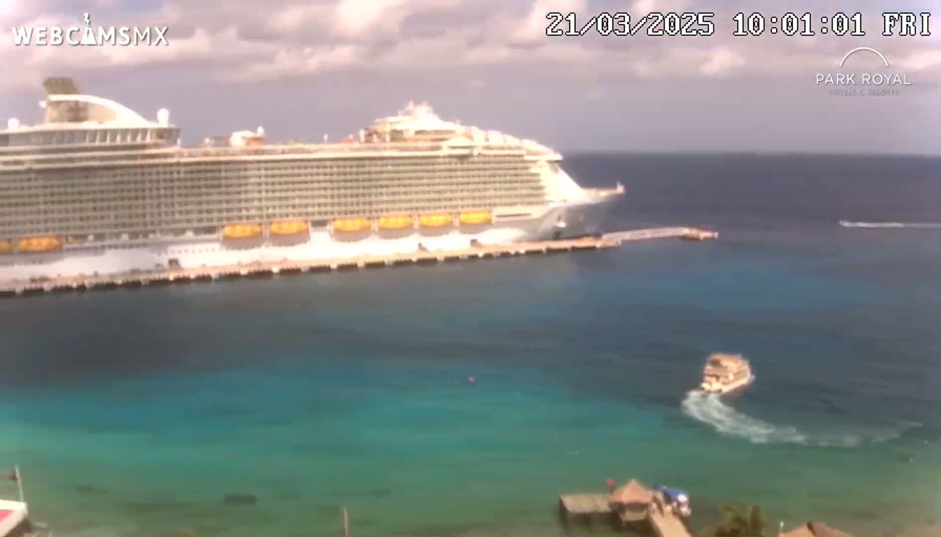 A large cruise ship is docked at a pier in calm, clear, turquoise water under a partly cloudy sky.