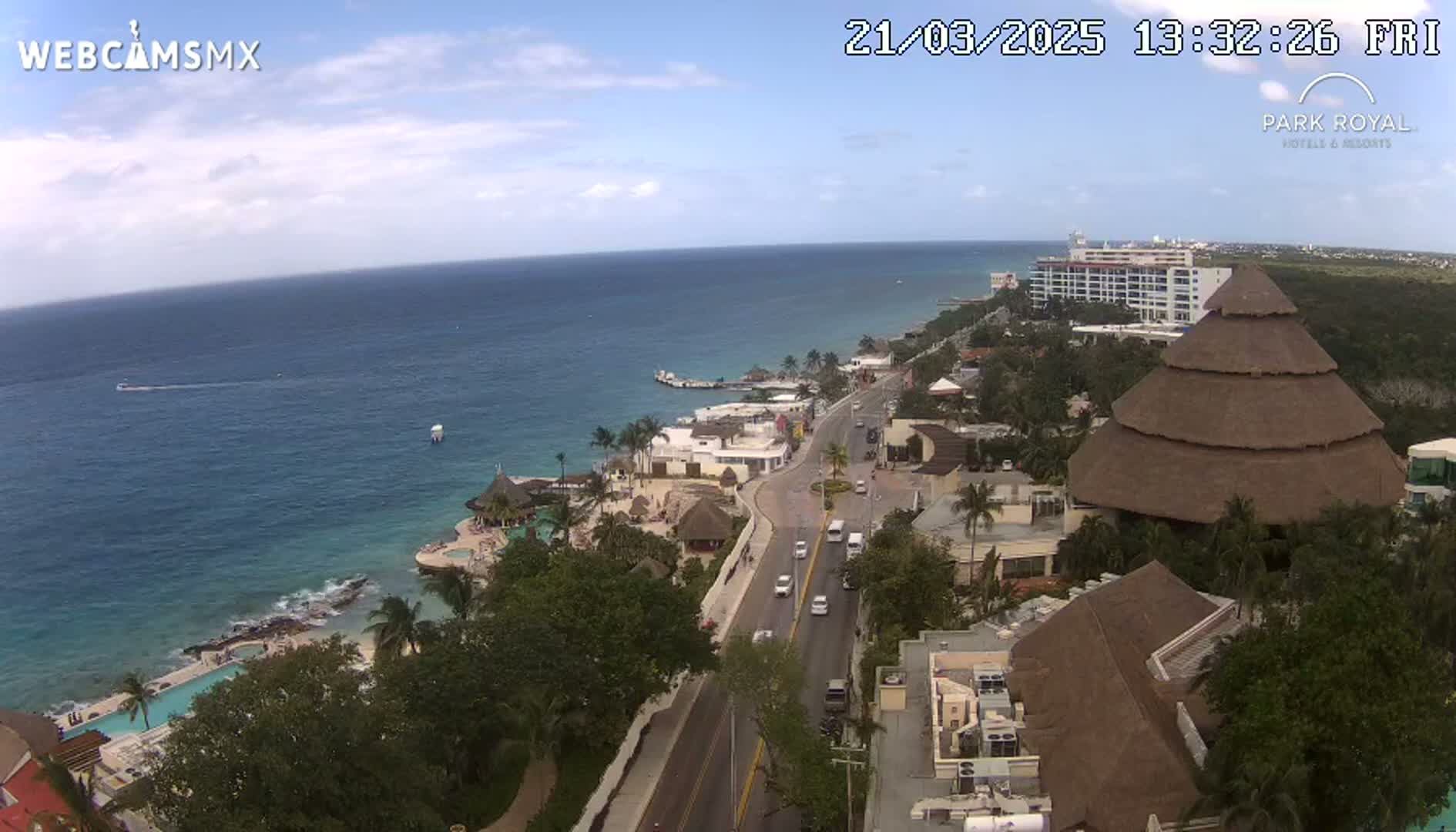 An aerial view shows a coastal town on a partly sunny day, with a calm ocean, a road leading to a resort with thatched-roof buildings, and several vehicles on the road.
