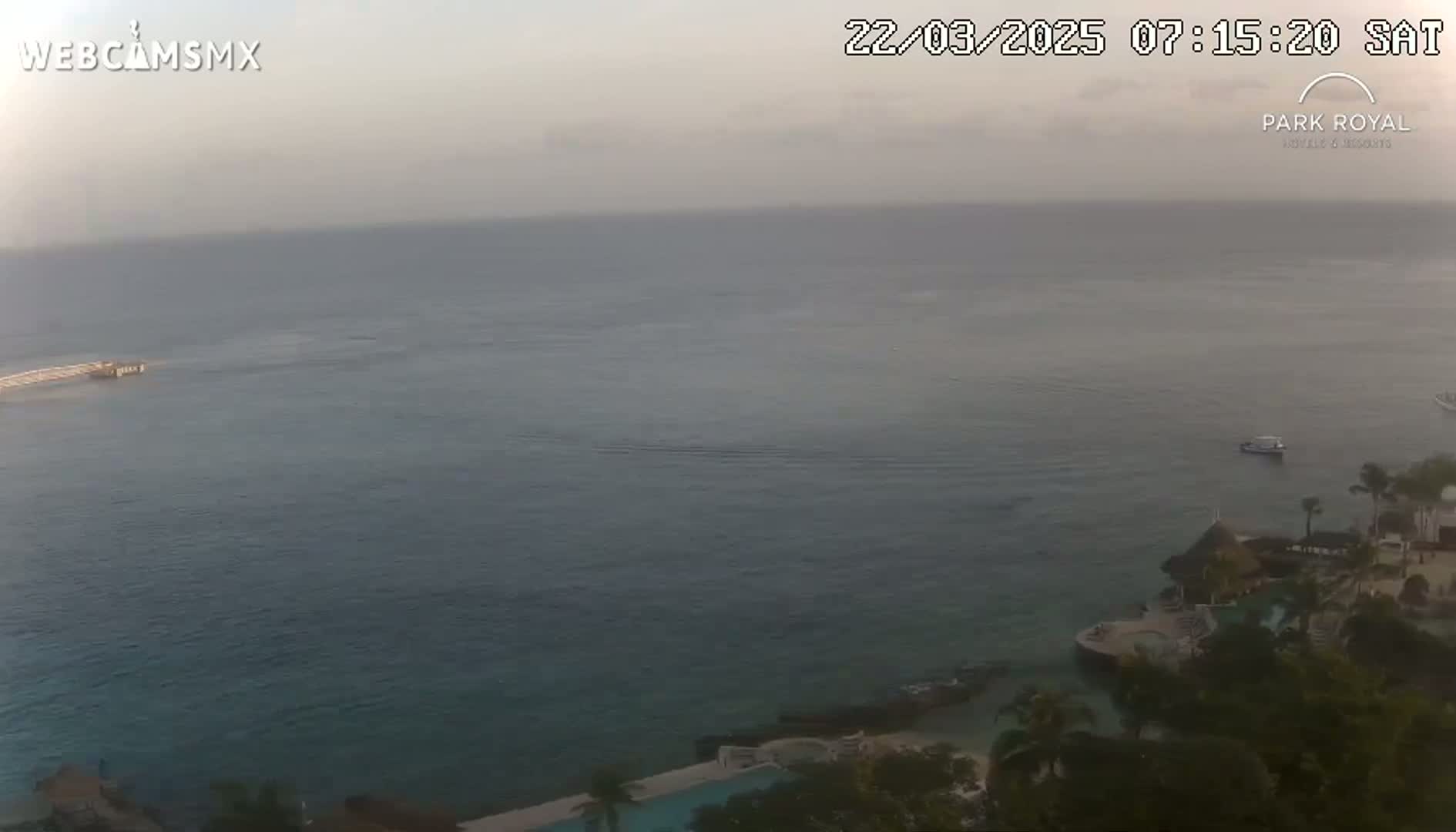 A hazy, calm ocean is seen from an elevated viewpoint, overlooking a resort with a pool and tropical vegetation.