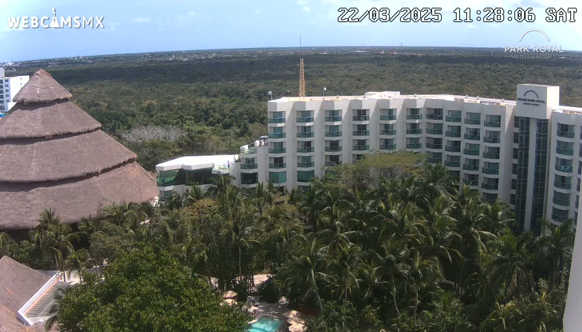 A large, multi-story white hotel is nestled within lush green tropical vegetation under a bright, sunny sky.