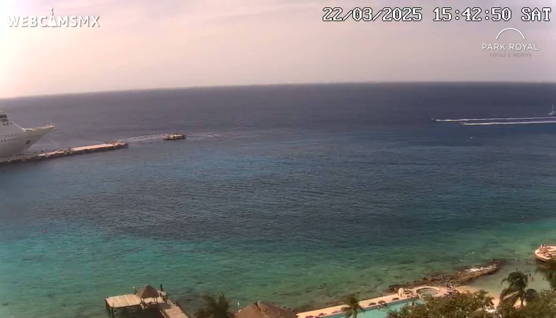 A large cruise ship is docked at a pier next to calm, clear turquoise water, with a smaller boat and a distant vessel visible on a sunny day.