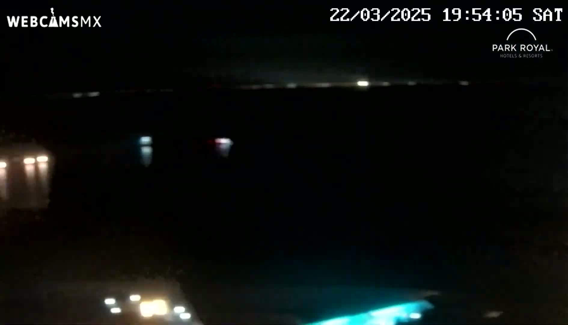 At night, a dark body of water is visible, with a few distant lights and a blurry, illuminated area in the bottom right corner.