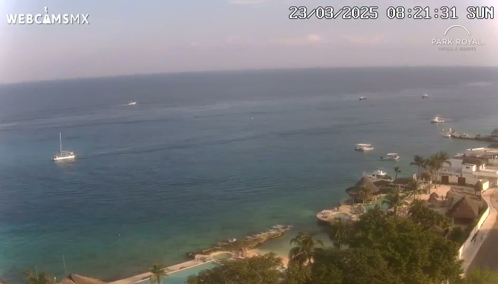 An aerial view shows a calm ocean with several boats and a resort on the coast on a hazy, sunny day.