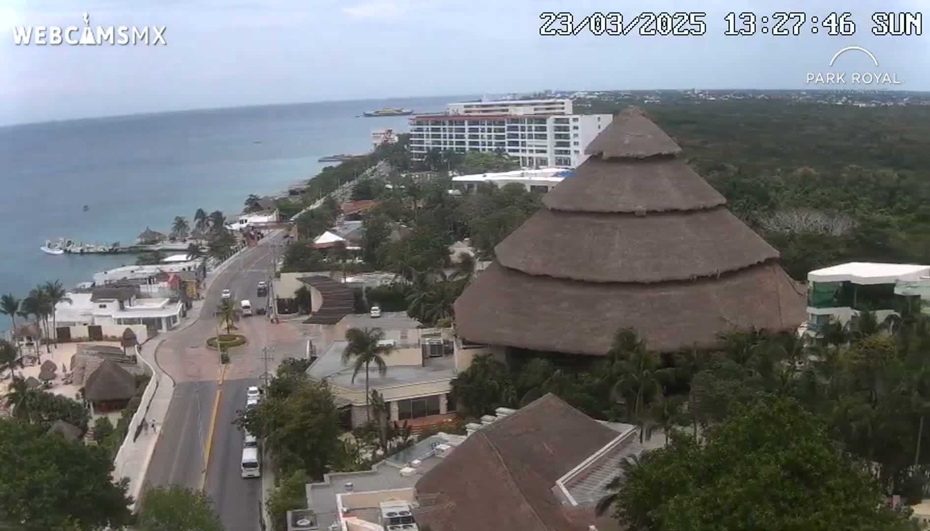 An aerial view shows a coastal resort town on a partly cloudy day, featuring a large thatched-roof building and a beachfront hotel.