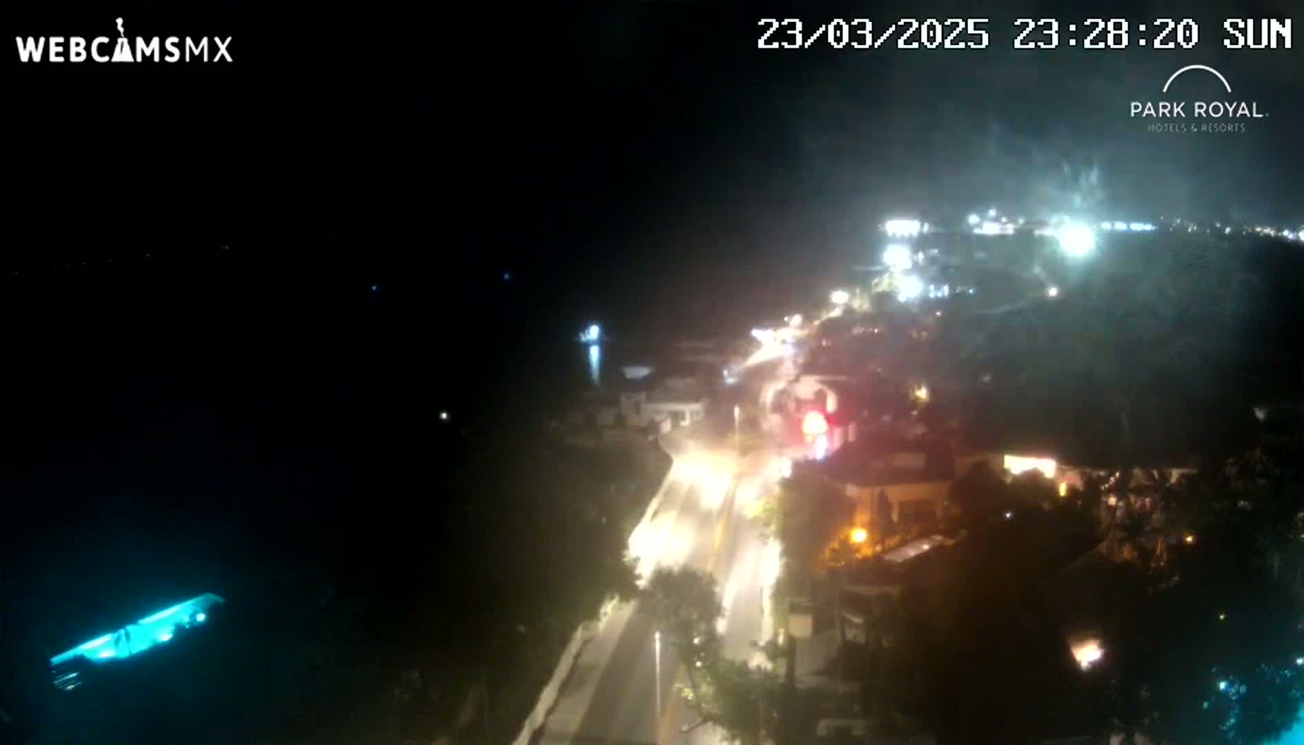 A nighttime, high-angle view shows a coastal road with vehicle lights and illuminated buildings, overlooking dark water.