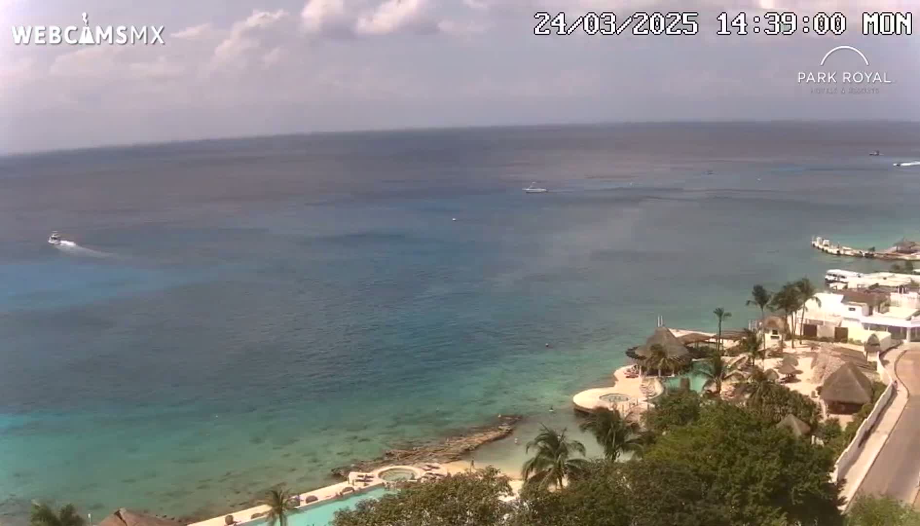 A partly sunny day shows a tropical resort on a rocky coastline with turquoise water, boats in the distance, and lush vegetation surrounding the resort's pools and buildings.