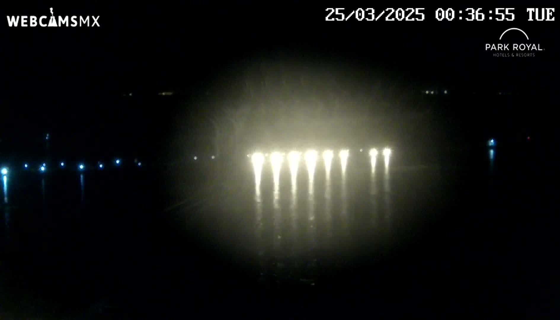 A nighttime, foggy view of a body of water reflecting bright lights from a distant structure.