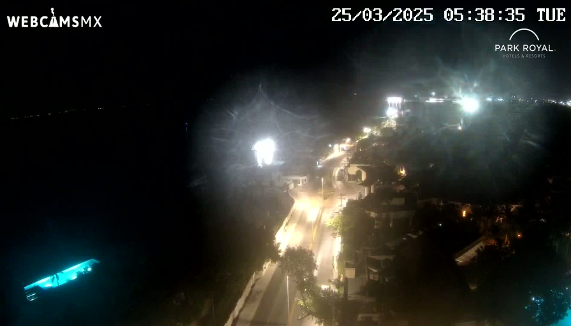 A nighttime, high-angle view of a coastal town with a brightly lit street and buildings, partially obscured by blowing spray or mist, near a body of dark water.
