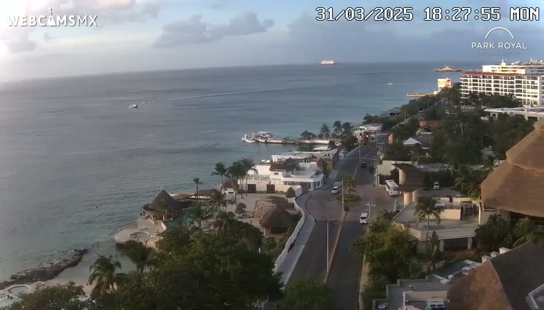 An aerial view shows a coastal resort town on a partly cloudy evening, with calm ocean waters and a large cruise ship in the distance.