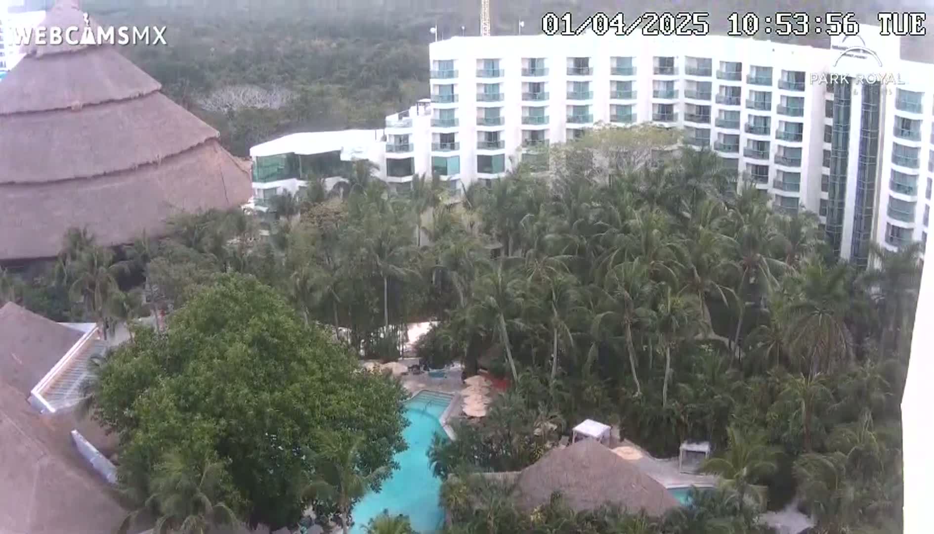 An aerial view shows a large hotel and resort complex nestled among lush tropical vegetation, including palm trees, with a swimming pool visible in the center.
