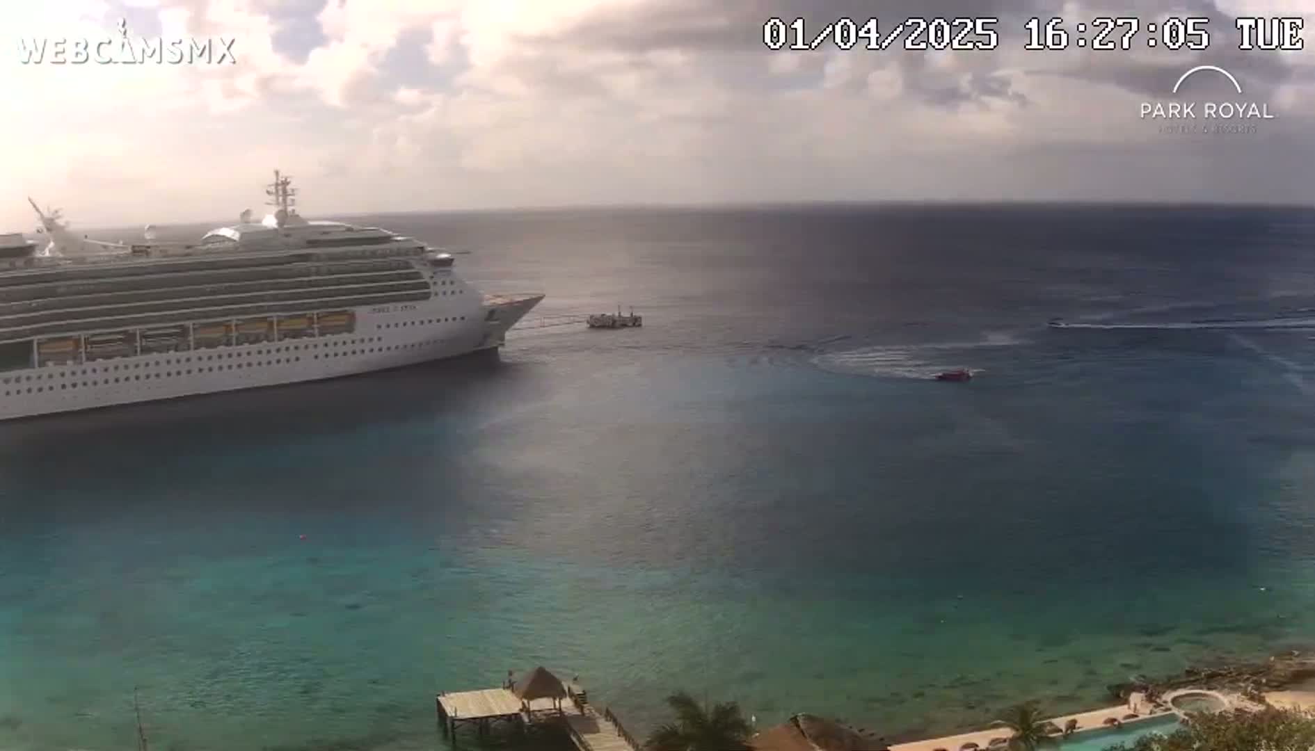 A large cruise ship is docked in calm, partly cloudy waters near a small dock and a resort.