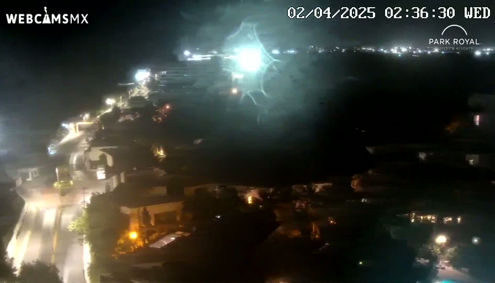 A nighttime aerial view shows a bright light display over a coastal town with illuminated roads and buildings.