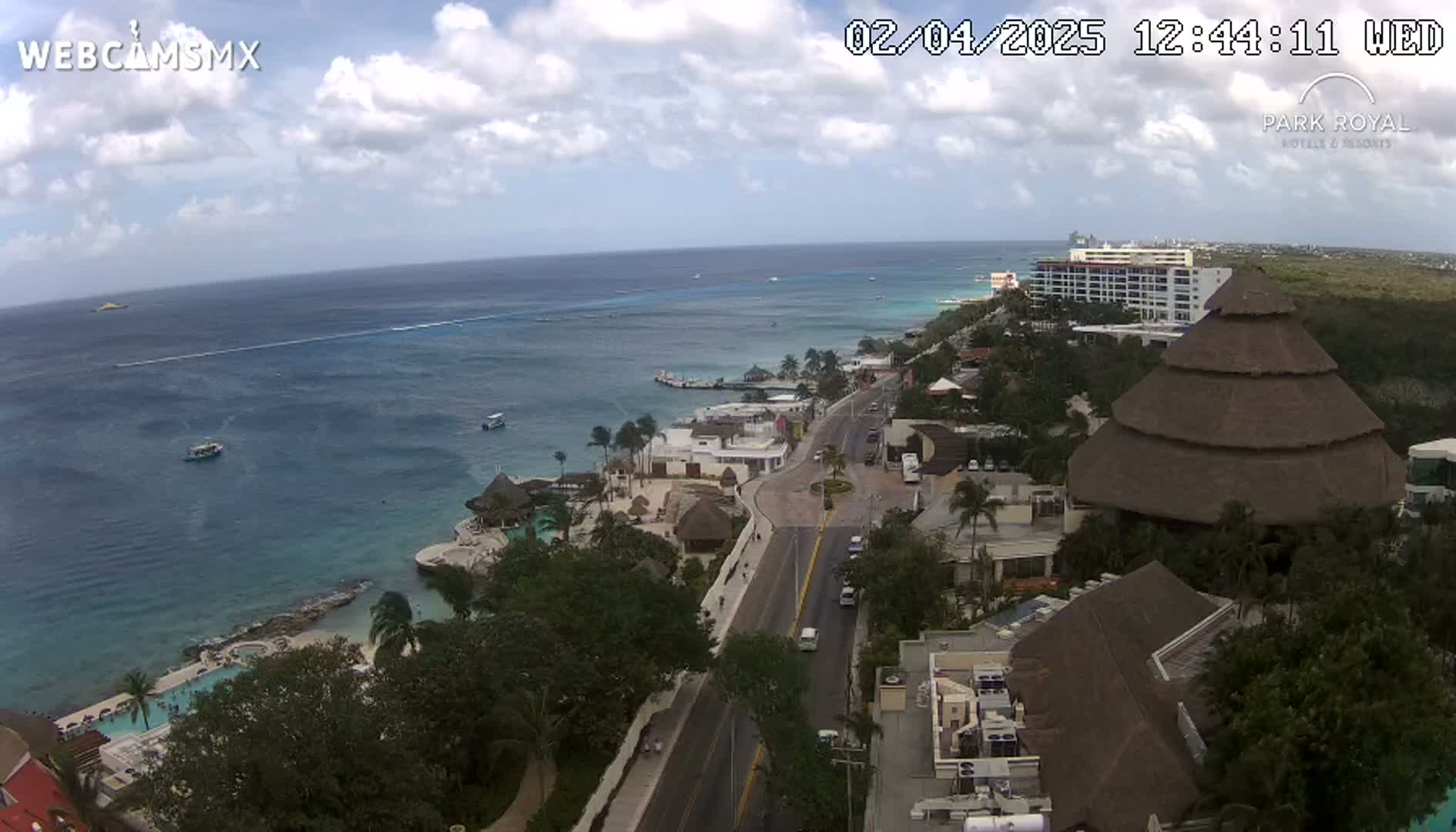 A partly cloudy day shows a coastal resort town with turquoise water, a road leading from the ocean to buildings, and several boats in the ocean.