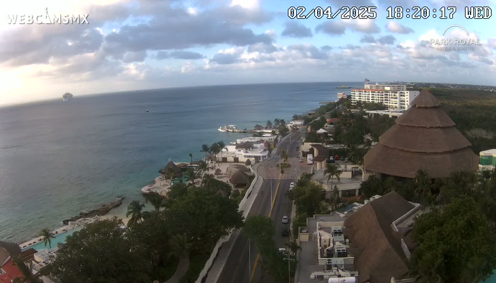 An aerial view shows a coastal resort town under partly cloudy skies, with a cruise ship visible in the distance.