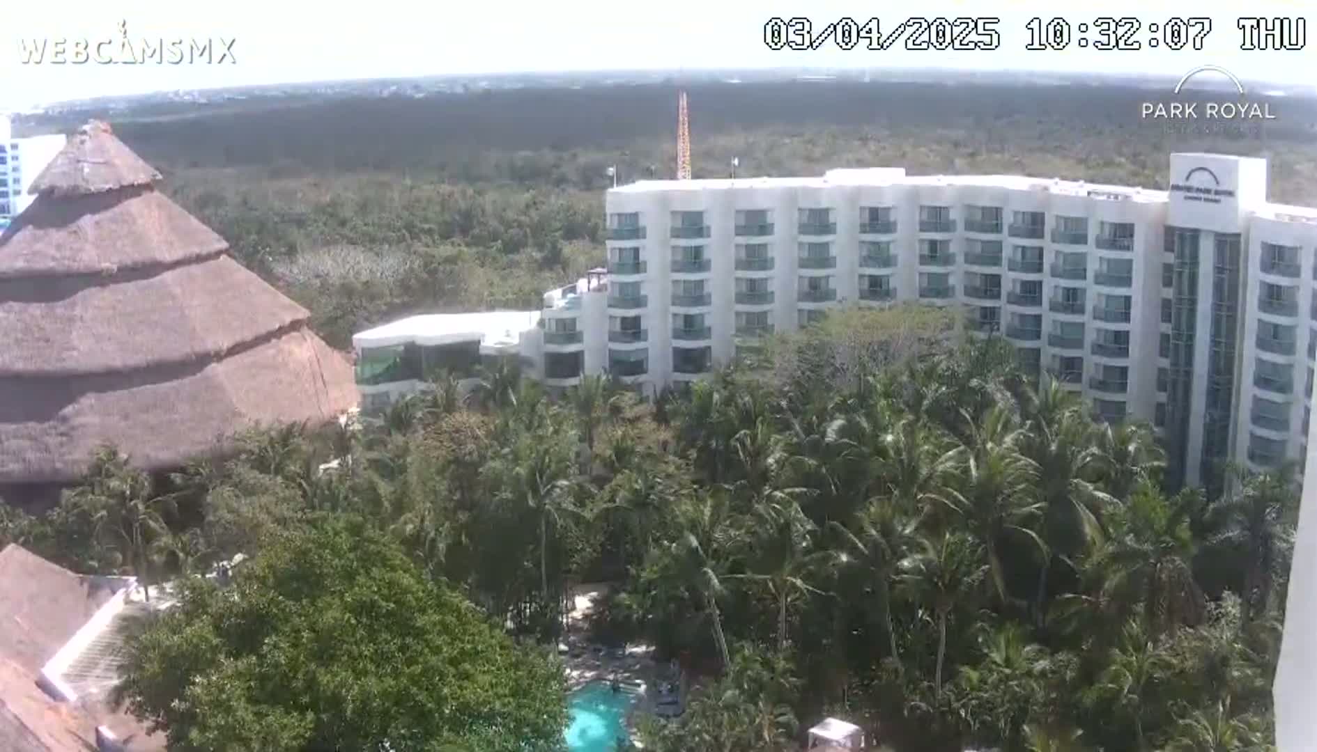 A large, white hotel is partially obscured by lush, tropical vegetation, including palm trees, with a thatched-roof structure visible in the foreground under a mostly clear sky.