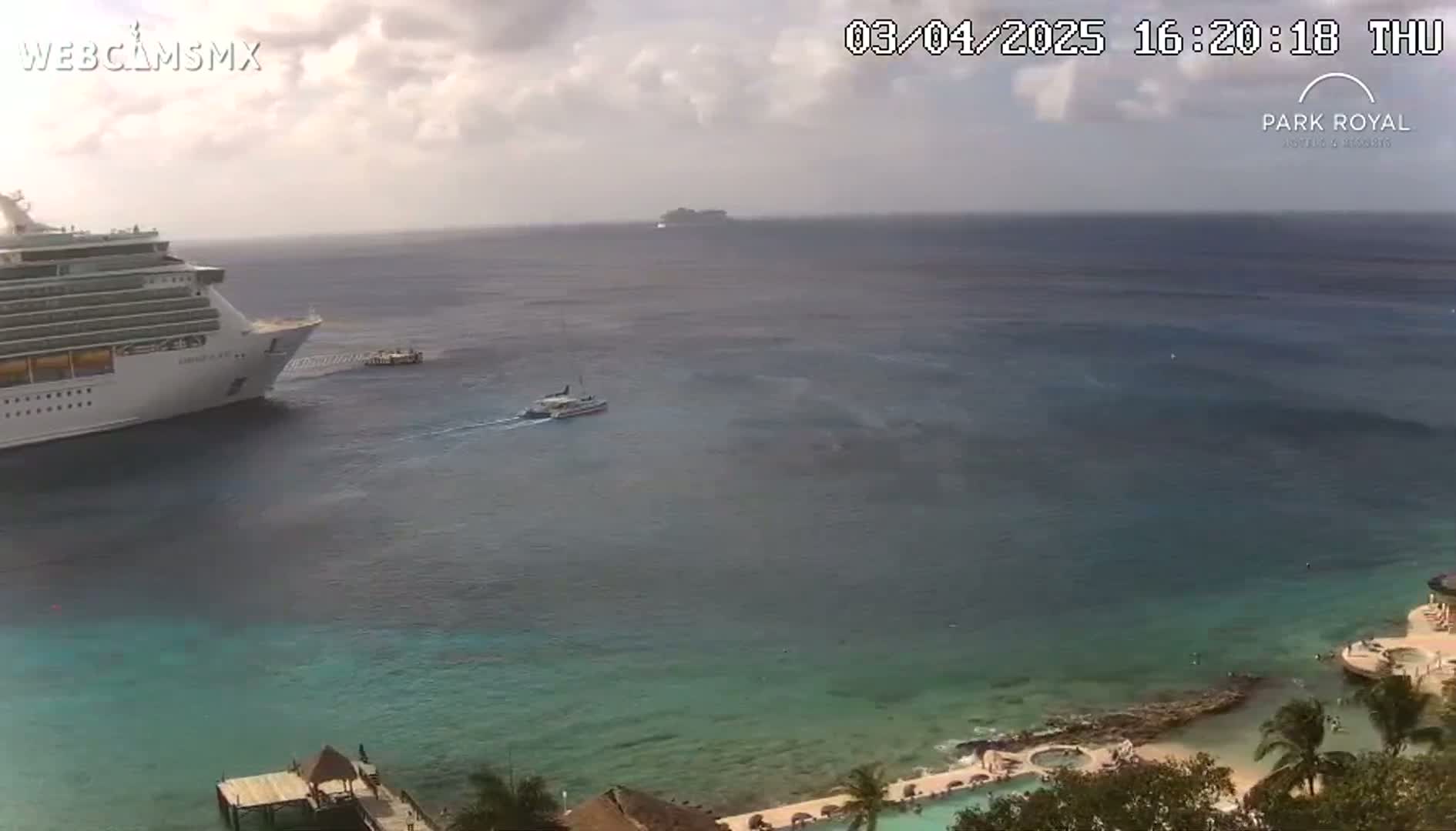 A large cruise ship is docked near a resort with clear, calm water under partly cloudy skies.