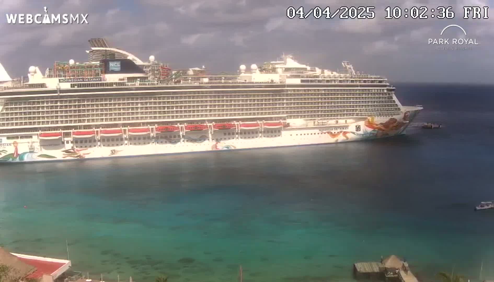 A large cruise ship is docked in clear, teal water under a partly cloudy sky.