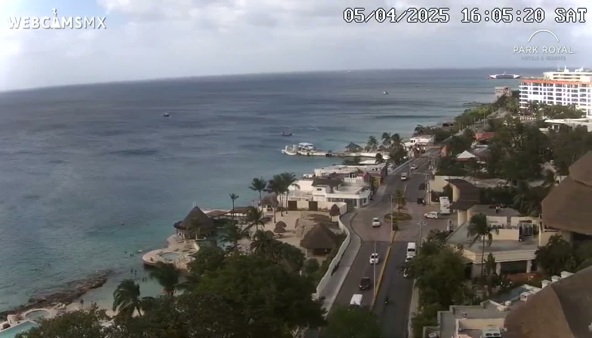 An aerial view shows a coastal resort town on a partly cloudy day, with calm ocean waters and a road winding through the buildings towards the shoreline.