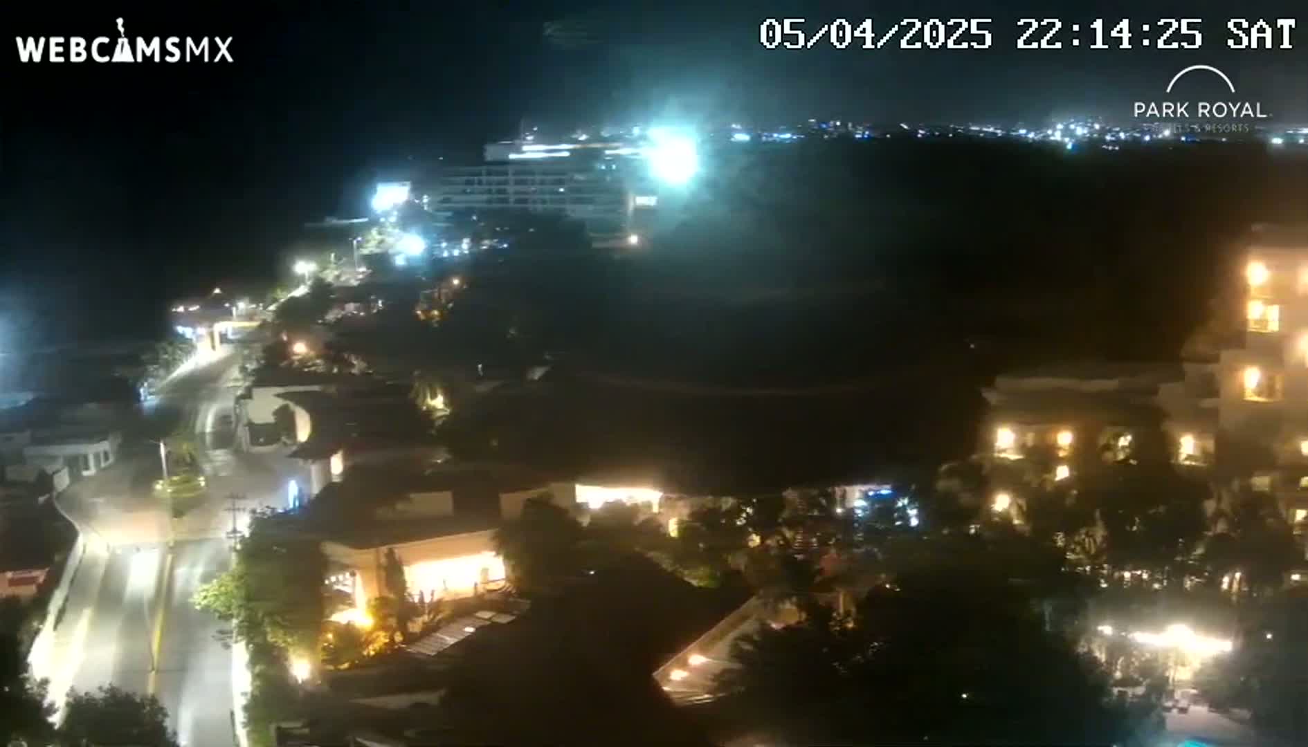 A nighttime aerial view shows a coastal town with illuminated buildings and roads, under a clear sky.