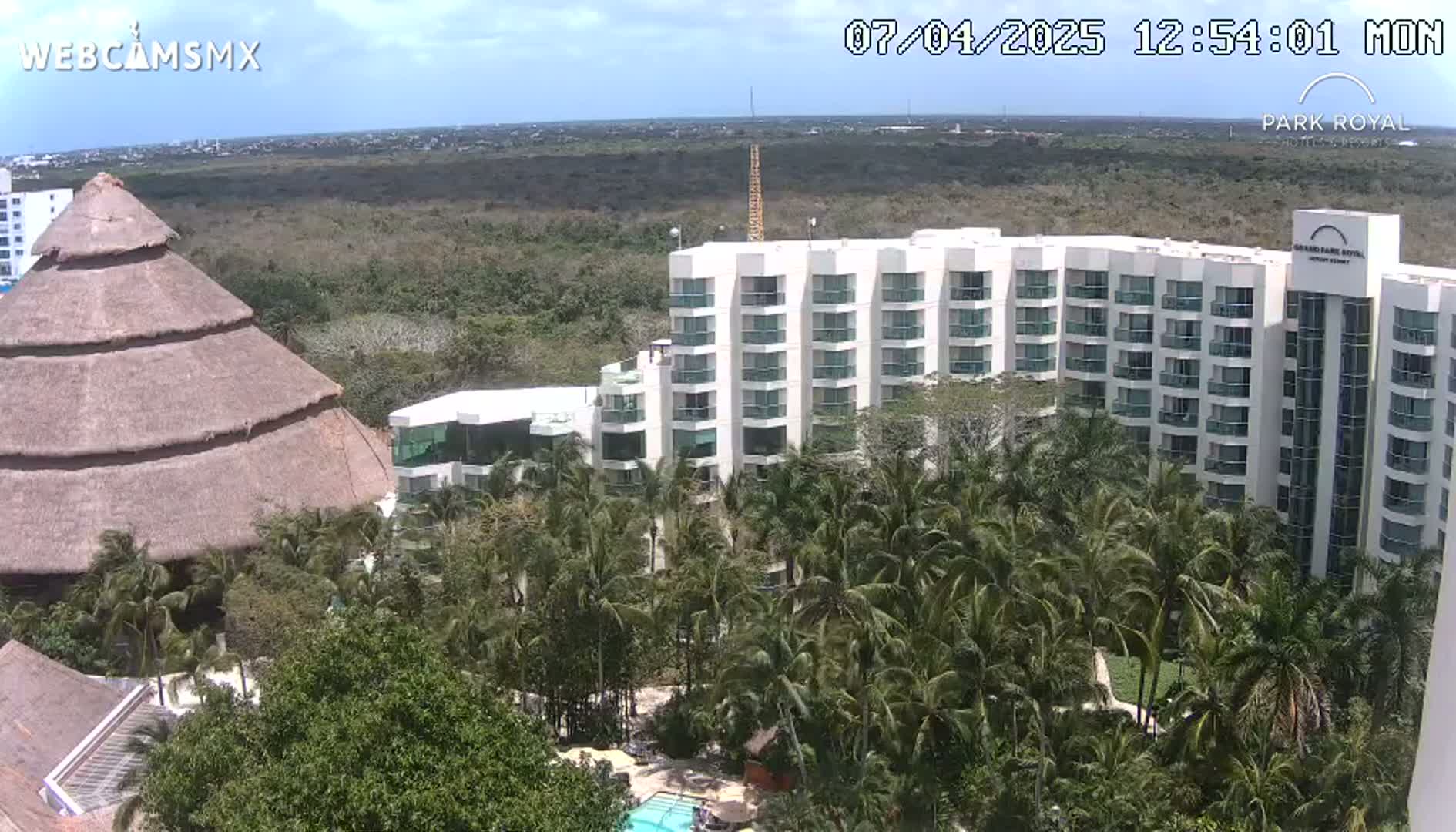A large, multi-story hotel complex with a thatched-roof building nearby, surrounded by lush palm trees under a partly sunny sky.